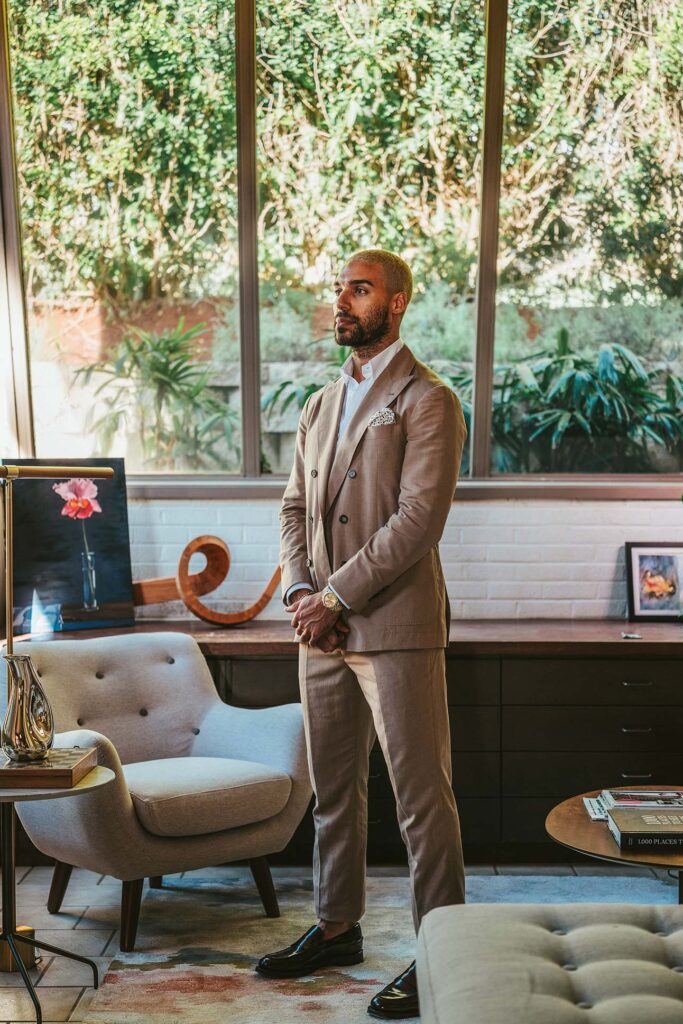 Man stands in a living room wearing a brown tailored suit that compliments his yellow gold luxury watch.