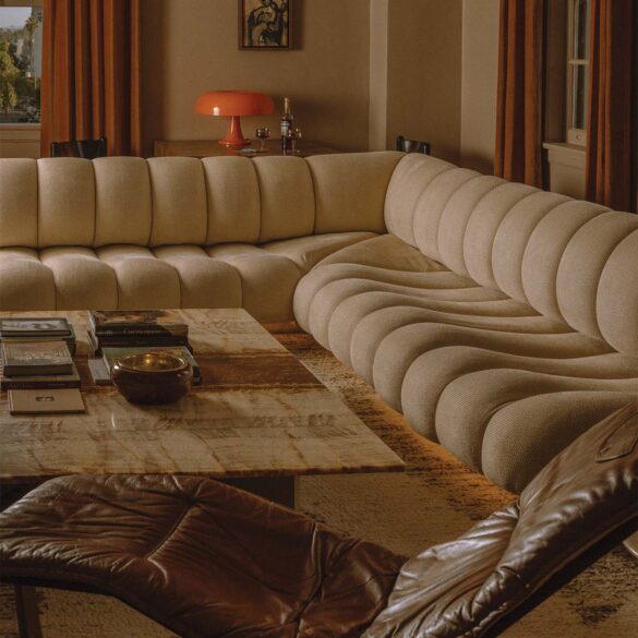A cozy living room with a beige, curved sectional sofa, a stone coffee table, and a brown leather lounger. Warm brown curtains cover the windows, and a red, retro-style lamp sits on a side table against the wall. The California design elements add laid-back elegance to the neutral-toned room.