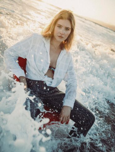 Woman seated partially in ocean waves, wearing a tied white shirt and dark pants with a serious expression. Sunlight illuminates her blonde hair and the surrounding water, creating a dramatic and serene scene. This fashion-forward moment showcases elegance amidst nature's beauty.