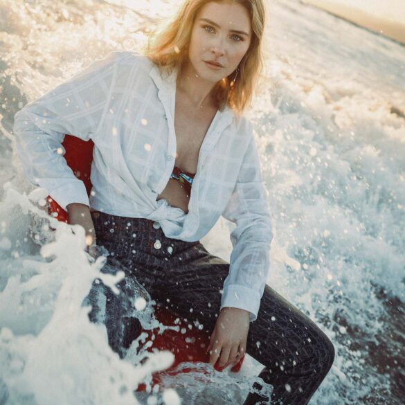 Woman seated partially in ocean waves, wearing a tied white shirt and dark pants with a serious expression. Sunlight illuminates her blonde hair and the surrounding water, creating a dramatic and serene scene. This fashion-forward moment showcases elegance amidst nature's beauty.